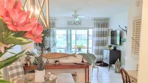 A spacious living room with a large window, a dining area with a wooden table, and a cozy seating arrangement. - “Cottage-Like” 2 Beds/2 Bath Condo Close To Beaches & The Heart Of ‘Old Stuart' (Stuart)