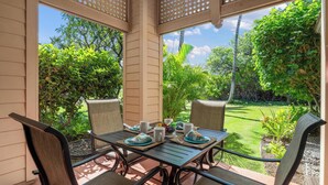A covered patio with a table set for two, surrounded by chairs, and a view of a lush garden. - Includes Mauna Lani Beach Pass.  101 Kulalani (Mauna Lani Resort)