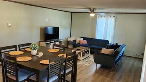 Een ruime woonkamer met een donker houten eettafel, stoelen en een bank. Een televisie is aan de muur bevestigd, en er is een plafondventilator te zien.