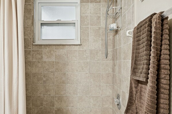 A bathroom with beige tiles, a showerhead, a towel rack, and a window with a frosted glass panel. - Lovely Lutz Living (Lutz)