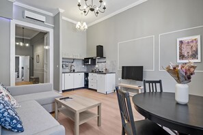 A modern living room with a grey sofa, a wooden coffee table, and a dining table with chairs. There is a kitchen area with white cabinets and a black range hood. A flat-screen TV is mounted on the wall, and a framed picture is displayed above the dining table. - Venus Apartments - Akacfa street I (Budapest)