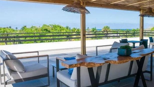 An outdoor seating area with a wooden table and chairs, a cushioned bench, and a view of the ocean. - Surf N Roll Performance Center (Cabarete)
