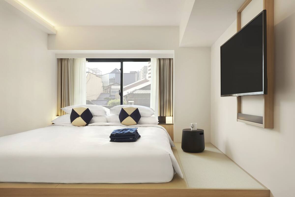 Ein Hotelzimmer mit einem Bett, einem Nachttisch, einem an der Wand befestigten Fernseher und einem Fenster mit Vorhängen.