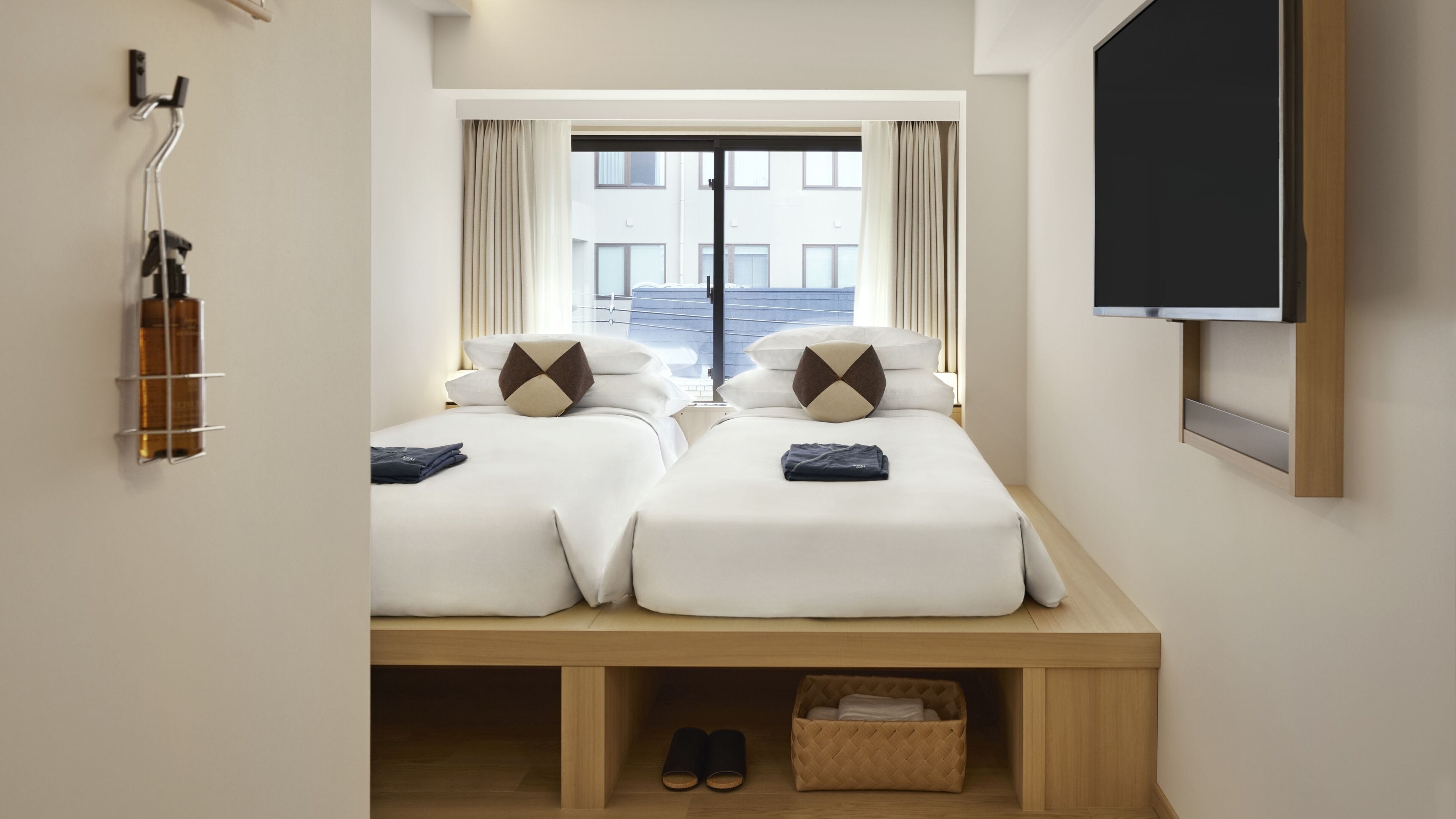 A hotel room with two beds, a TV, a window with curtains, a shoe rack, and a small storage area.