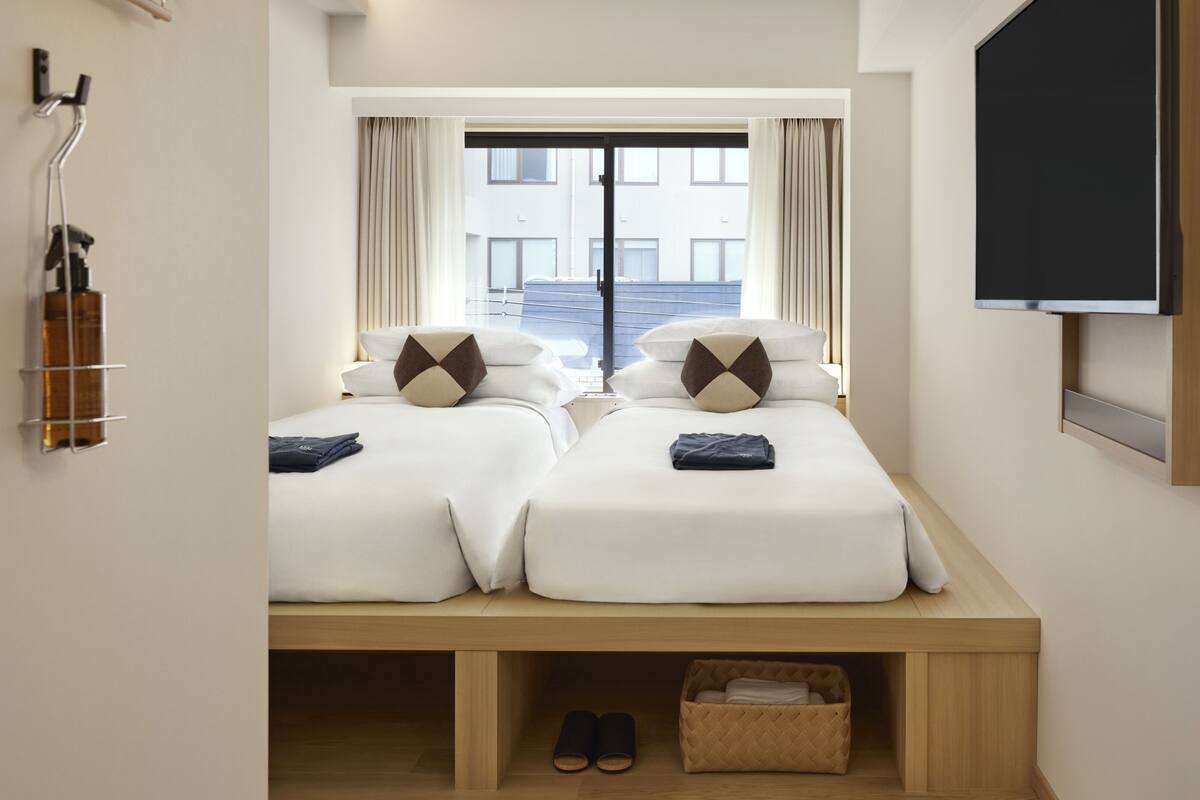 Ein Hotelzimmer mit zwei Betten, einem Fernseher, einem Fenster mit Vorhängen, einem Schuhregal und einer kleinen Ablage.
