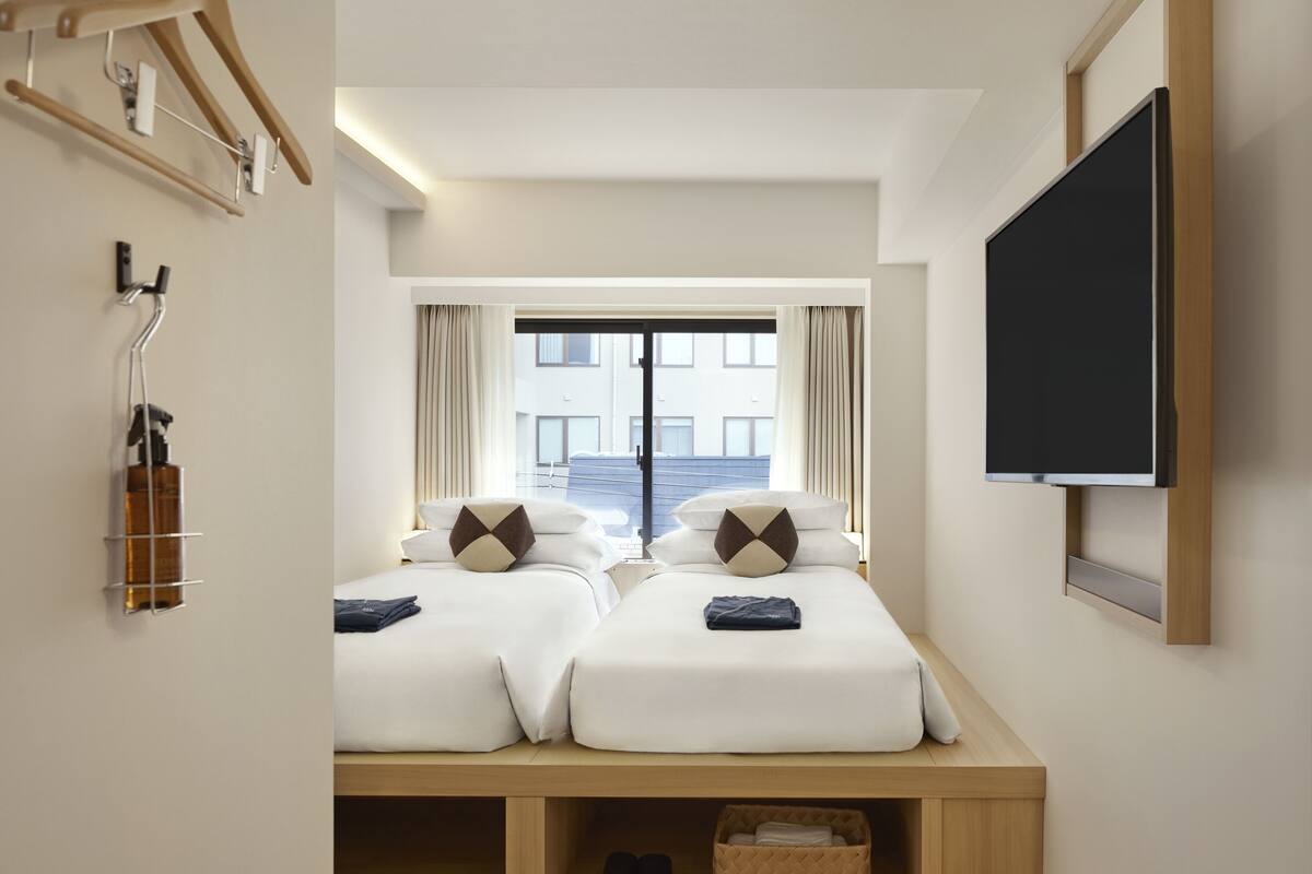 Ein Hotelzimmer mit zwei Betten, einem Fernseher, einem Fenster mit Vorhängen und einem Schuhregal.