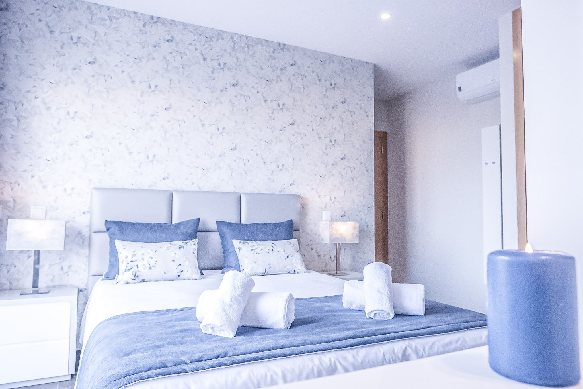 Une chambre d’hôtel avec un lit, deux tables de chevet, une petite table et une bougie bleue.