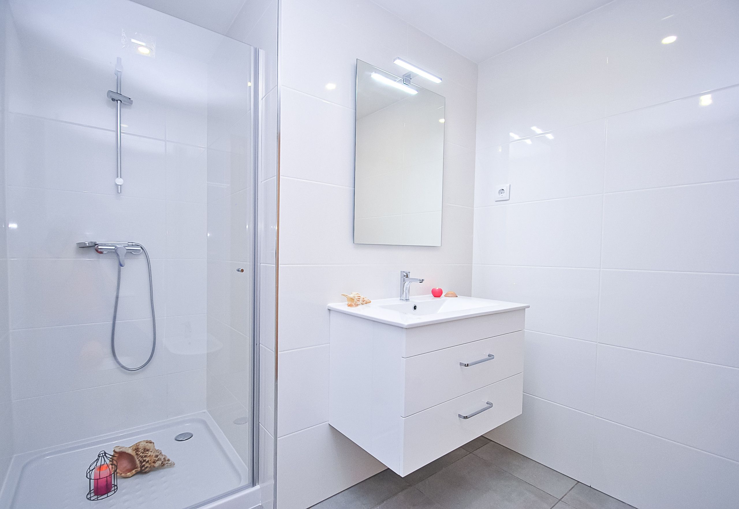 Un baño moderno con cabina de ducha de vidrio, un lavabo blanco con un espejo arriba y suelo de baldosas.