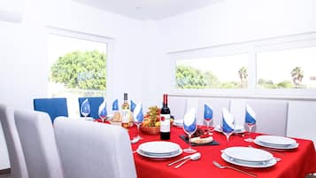 A dining area with a red tablecloth, white plates, and blue napkins. There is a bottle of wine and a basket of fruit on the table.