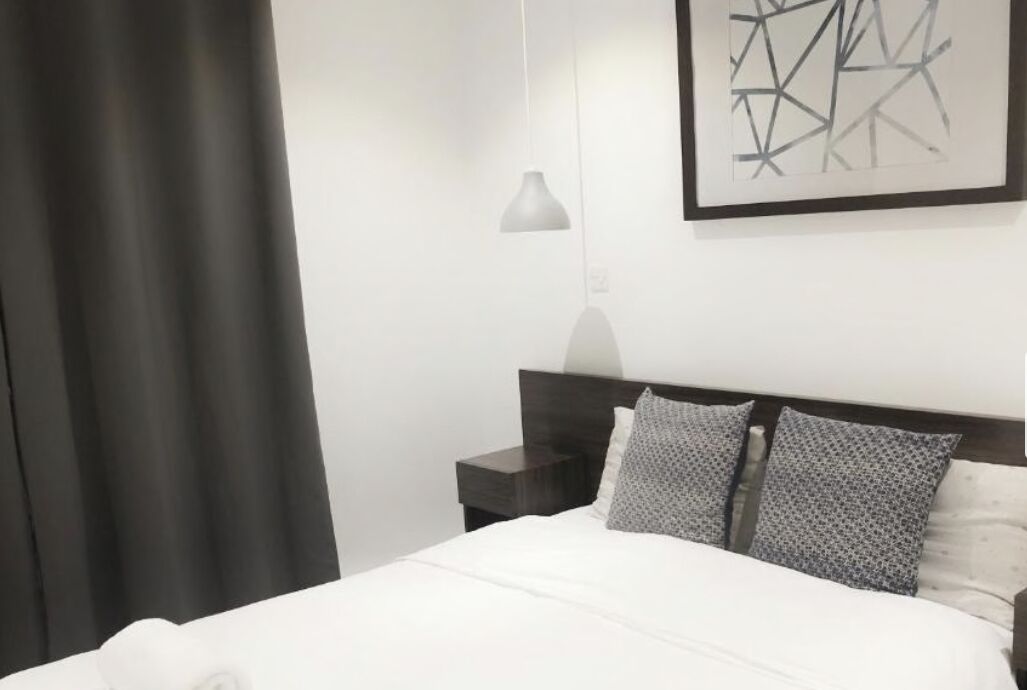 Una cama bien hecha con edredón blanco y dos almohadas estampadas, una mesita de noche con una lámpara y una obra de arte abstracta enmarcada en la pared.