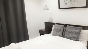 Un lit bien fait, avec une couette blanche et deux coussins à motifs, une table de chevet avec une lampe et une œuvre d’art abstraite encadrée accrochée au mur.