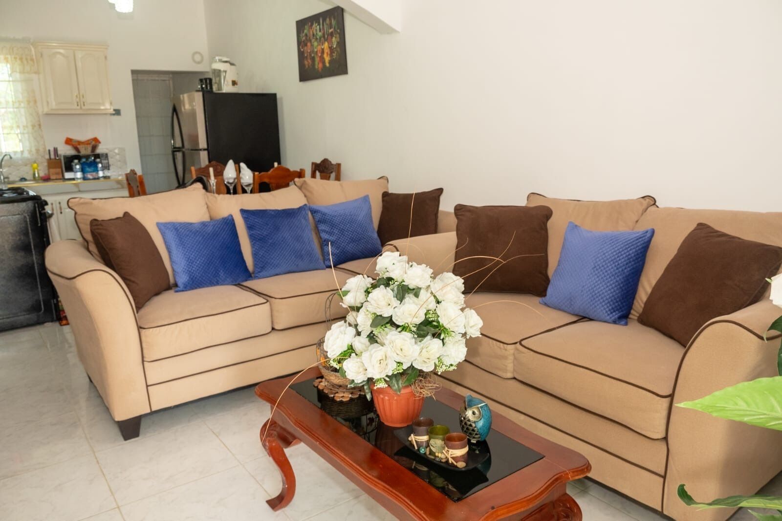 Ein Wohnzimmer mit einem beigen Sofa, blauen und braunen Kissen, einem Couchtisch mit Blumen und einer Küche im Hintergrund.