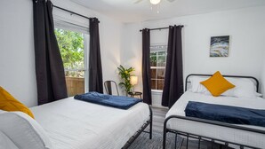 A bedroom with two beds, a ceiling fan, a window with curtains, a small table with a lamp, and a plant.