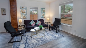 A living room with a dark-colored sofa, two black chairs, a small round coffee table, and a blue patterned rug. There are three wall-mounted lamps and two floral paintings on the walls.