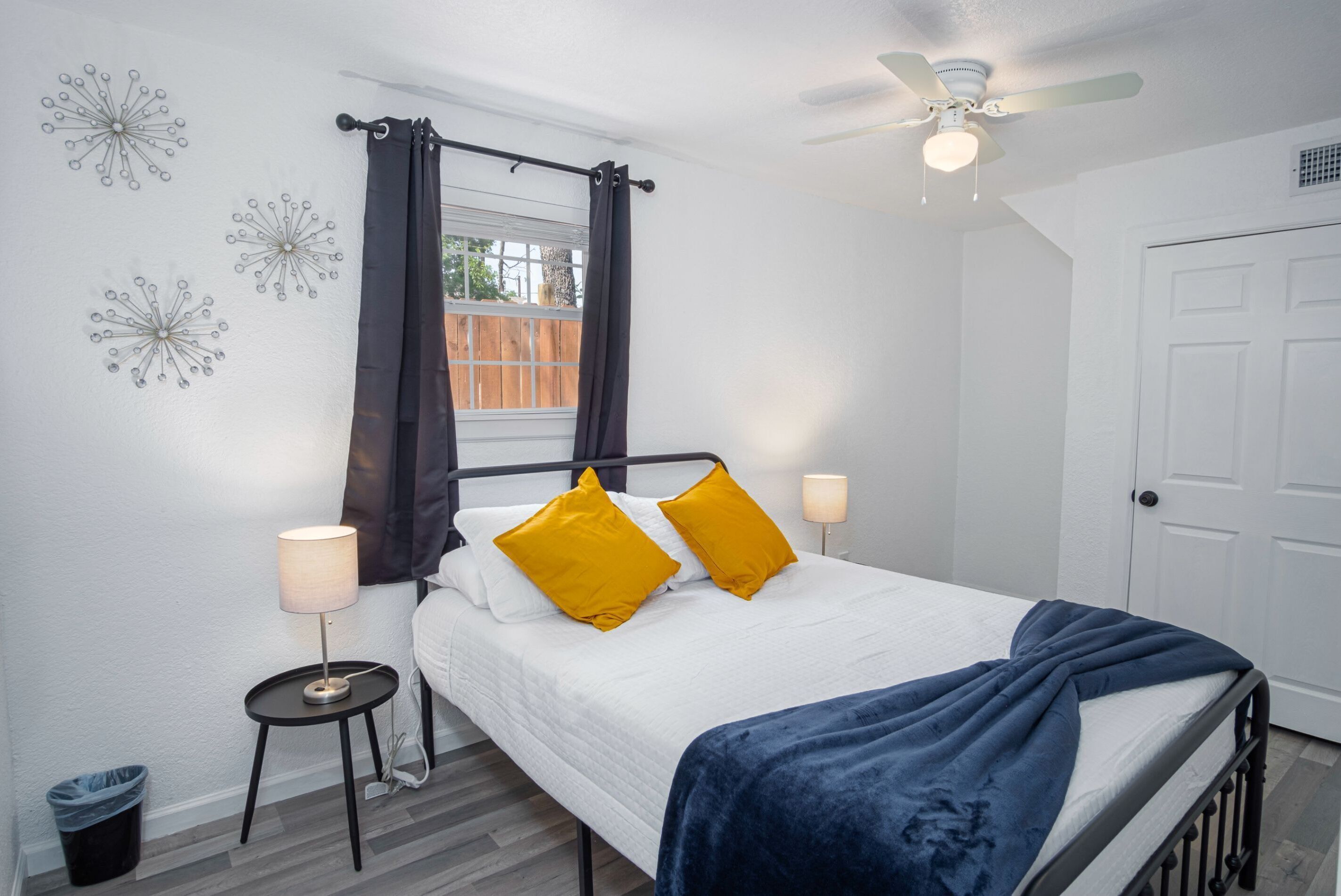 Ein Schlafzimmer mit Bett, Deckenventilator, Fenster mit Vorhängen, Nachttischlampe und Mülleimer.