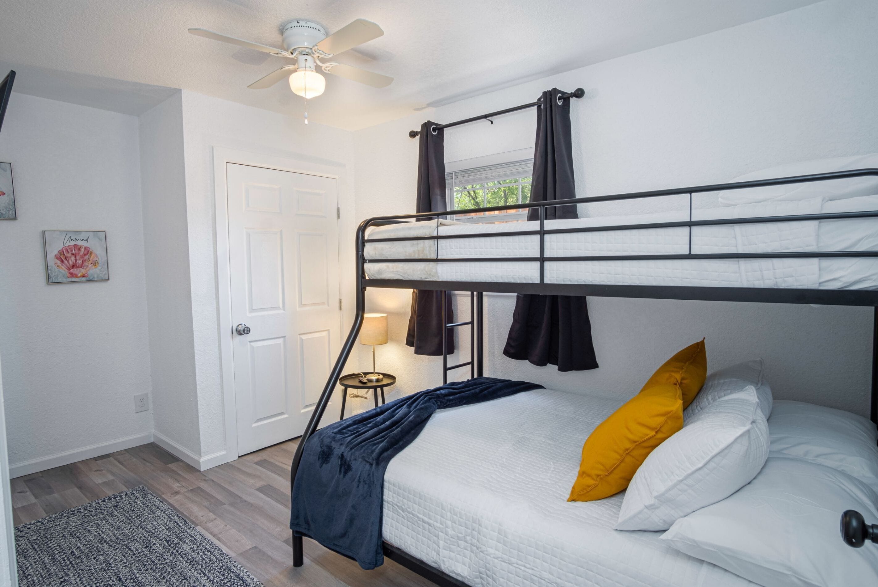 A bunk bed room with a ceiling fan, a door, a small table with a lamp, and a window with curtains.