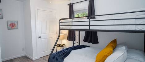 A bunk bed room with a ceiling fan, a door, a small table with a lamp, and a window with curtains.