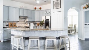 A modern kitchen with light blue cabinets, a large island with bar stools, and a dining area visible through an archway. - Palm Coast Oasis Retreat- 15 min to beach (Palm Coast)