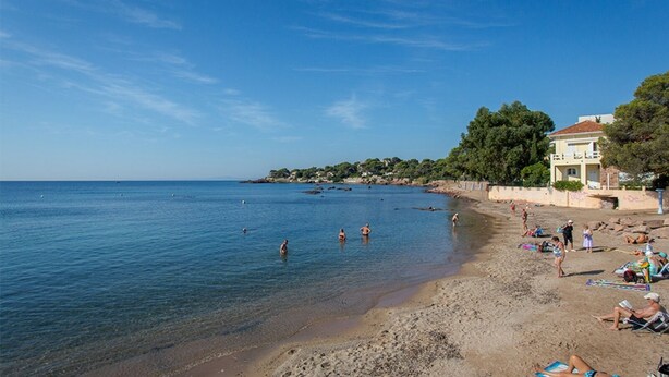 Una spiaggia con acque cristalline, persone che nuotano e si rilassano al sole, e un edificio con un balcone sulla destra.