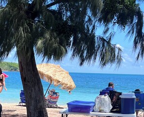 Escena de playa con una sombrilla de paja, sillas de playa y una hielera sobre la arena.