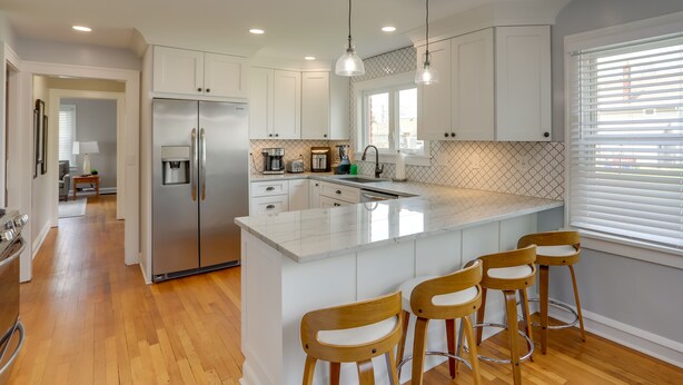 A modern kitchen with white cabinets, a stainless steel refrigerator, a marble island with bar stools, and pendant lighting. - Walk to Lake Erie: Chic Home, Private Yard & Deck (Avon Lake)
