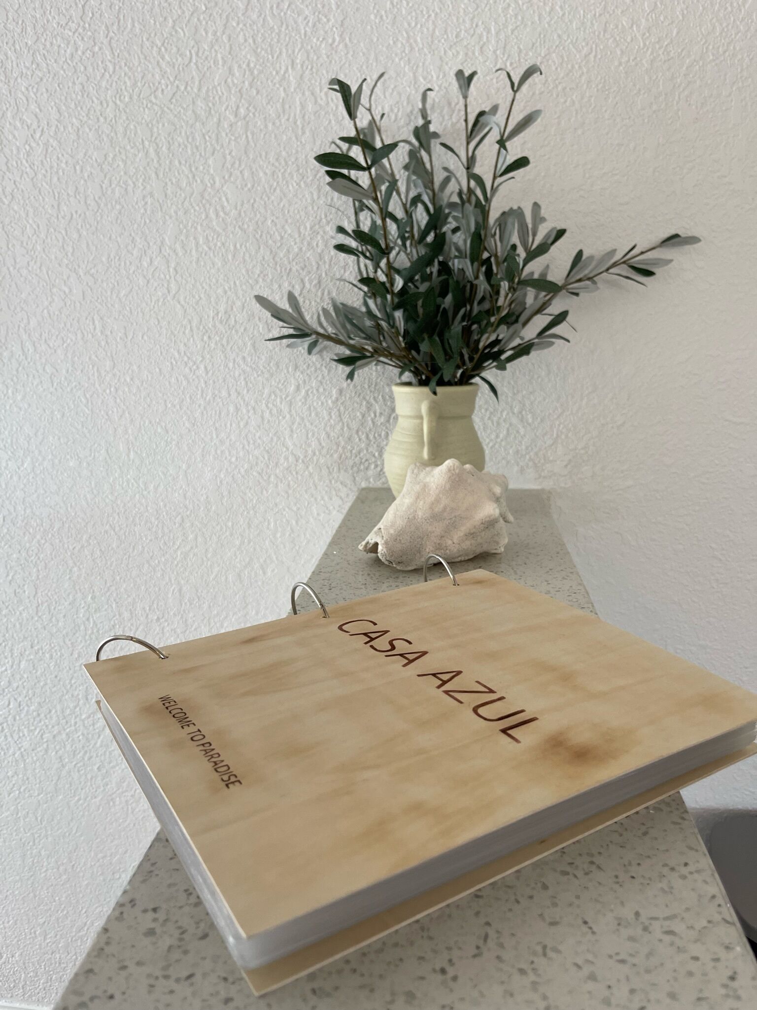 Un livre en bois intitulé « CASA AZUL », portant l’inscription « BIENVENUE AU PARADIS », est posé sur un mur blanc à texture, à côté d’un vase contenant des feuilles vertes.