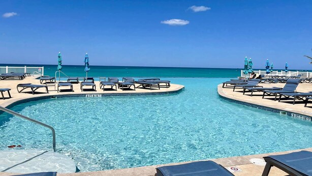 A pool area with lounge chairs and umbrellas, clear blue water, and a view of the ocean. - Casa Azul welcomes you to a stunning ocean view condo steps away from the beach (Bailey Town)