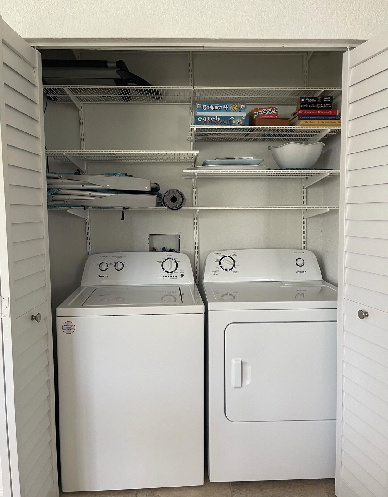A laundry room with two front-loading washing machines, a drying rack, and shelves with books and bowls.