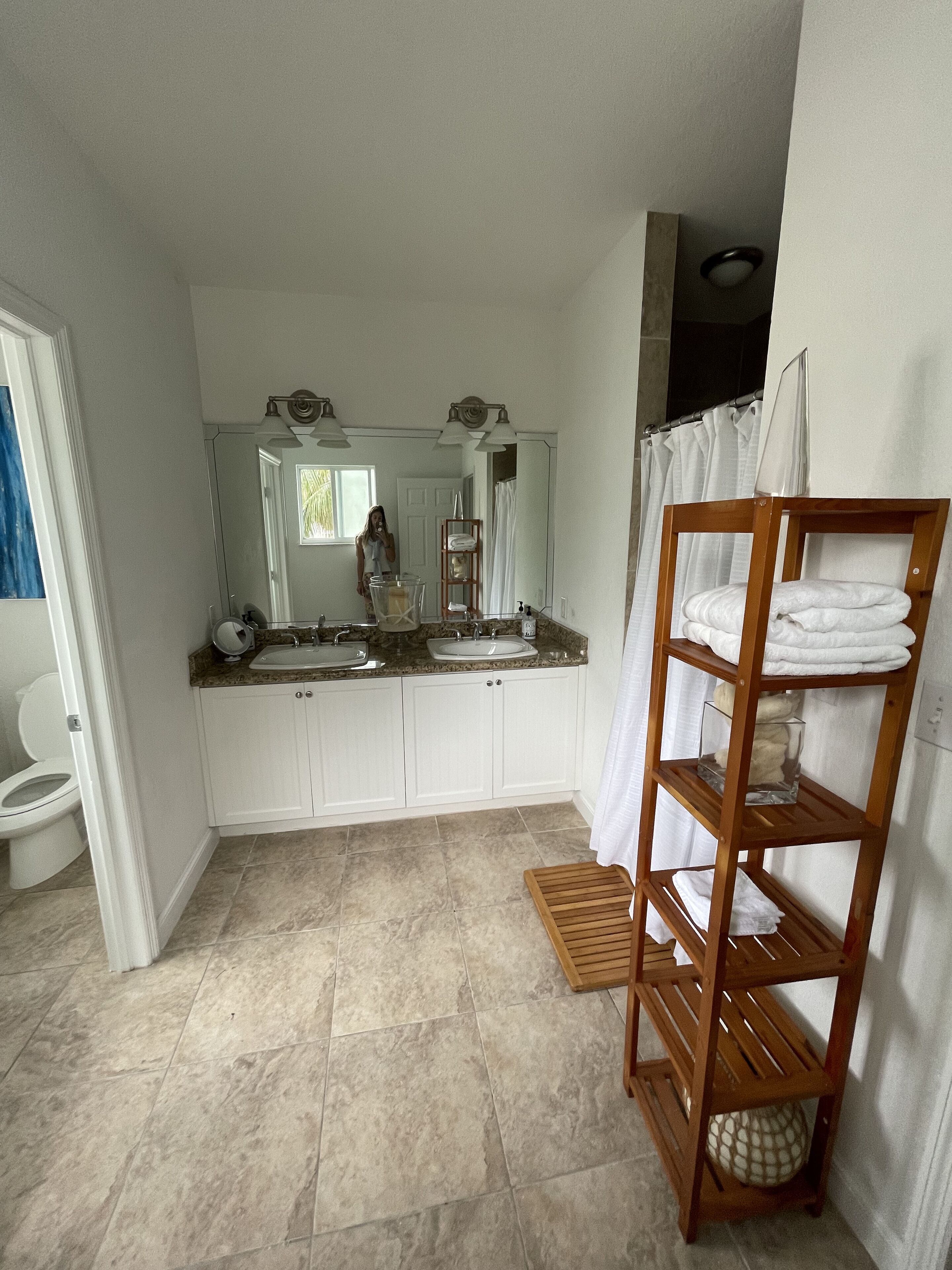 A bathroom with a double sink, a large mirror, a wooden shelf with towels, and a shower curtain.