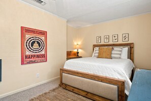 Quarto com cama, luminária na parede, pôster emoldurado e uma pequena mesa.