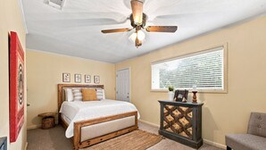 Une chambre à coucher comprenant un lit, une tête de lit en bois, un ventilateur de plafond, une fenêtre avec des stores et une petite armoire.
