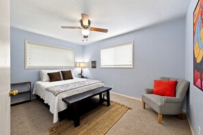 Une chambre à coucher comprenant un lit, un banc, une chaise, un ventilateur de plafond et deux fenêtres équipées de stores.