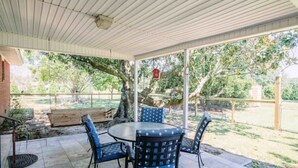 A covered patio area with a round table and four chairs, surrounded by a wooden fence and trees.