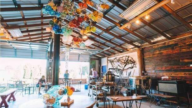 An industrial-style interior with exposed beams, a rustic brick wall, and a variety of colorful glass objects hanging from the ceiling. - Historic FarmHouse with Goats & Glass Blowing (Hattiesburg)