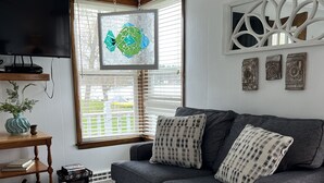 A living room with a grey sofa, two patterned pillows, a woven coffee table, a flat-screen TV, a decorative mirror, and a window with blinds. - Keeney Kottage (Six Lakes)