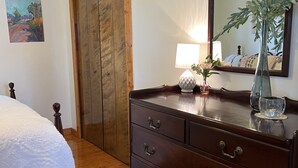 A bedroom with a wooden dresser, a bed with a white duvet, a painting on the wall, and a mirror reflecting a plant and a lamp. - Keeney Kottage (Six Lakes)