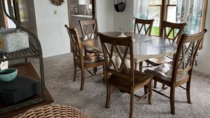 A dining area with a round table and wooden chairs, a chandelier, and a view of a garden through large windows. - Keeney Kottage (Six Lakes)