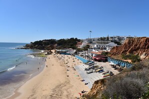 Uma praia costeira com uma fileira de cabanas azuis, areal arenoso e edifícios ao fundo.