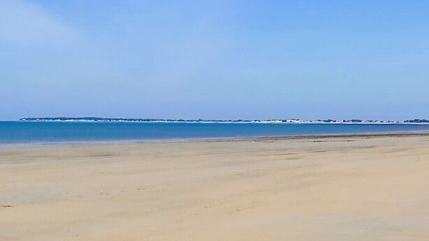 Una playa extensa de arena clara con cielo despejado y una costa lejana.