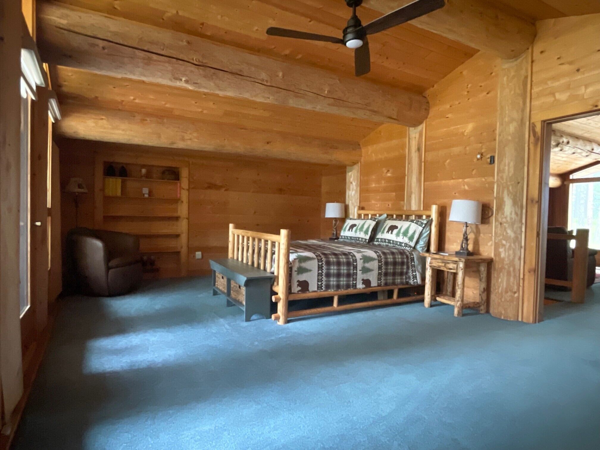 A wooden cabin bedroom with a bed, two bedside tables, a chair, a ceiling fan, and a window.