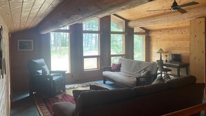 Interior acogedor de una cabaña con techo de madera, un sofá, una silla, una mesita con una lámpara y un televisor.
