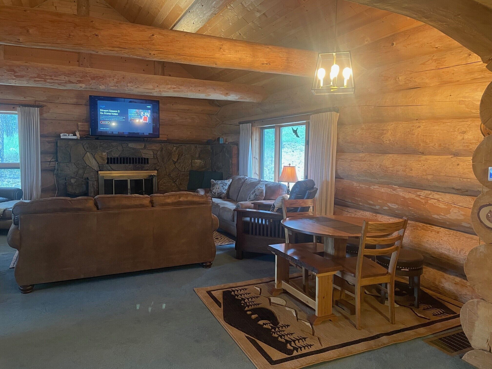 A cozy log cabin interior with a stone fireplace, a television mounted on the wall, a wooden dining table with chairs, and a sofa.