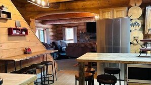 Interior rústico de una cabaña con una zona de comedor de madera, taburetes de bar y una cocina con un refrigerador grande.