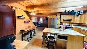 Interior rústico de una cabaña con barra de madera, cocina con ollas colgantes y un espacio de estar acogedor.