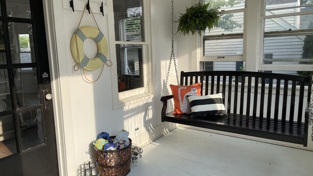 A porch with a black swing, a wicker basket, and a life preserver on the wall.