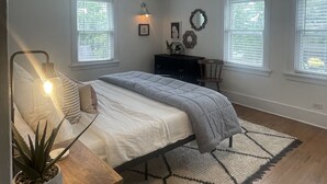 A bedroom with a bed, bedside tables, a desk, a chair, a rug, and two windows with blinds.