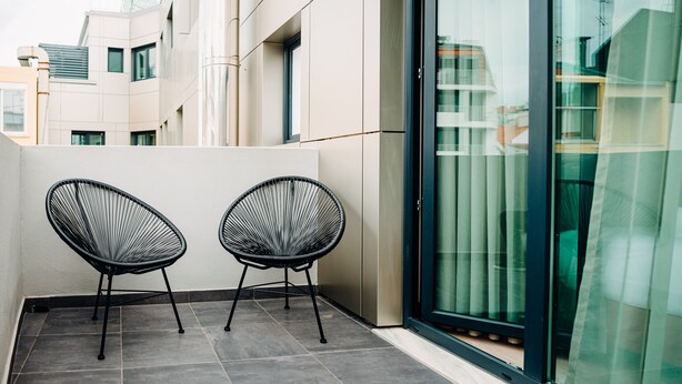 A balcony with two black wire-framed chairs, a tiled floor, and a glass door leading to a building with a modern design. - Next Level Premium Hotels (Lisbon)