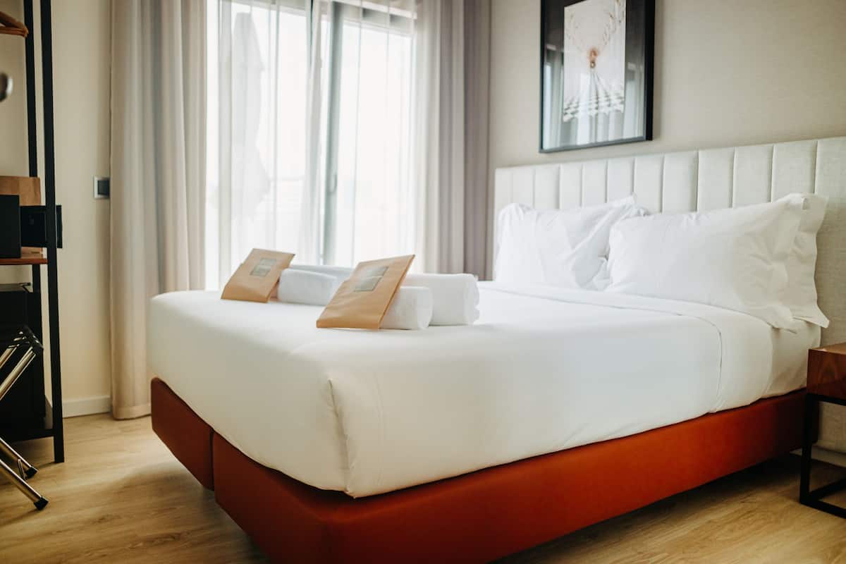 A hotel room with a large bed, a wooden nightstand, a framed picture on the wall, and a window with curtains.