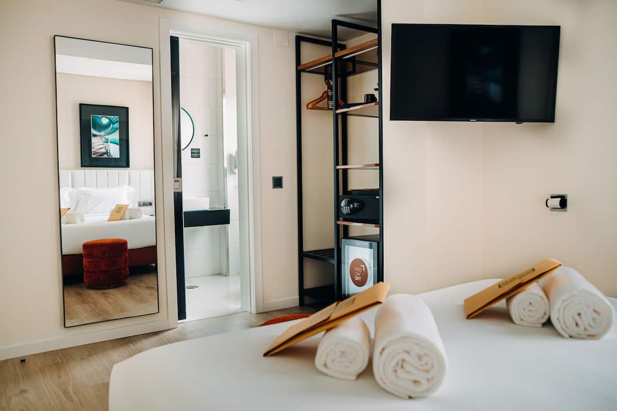 A hotel room with a bed, a TV mounted on the wall, a wardrobe, and a mirror reflecting the room.