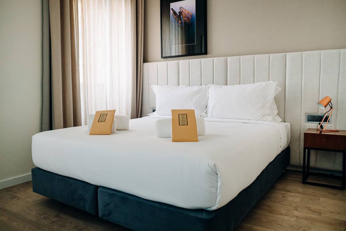 A neatly made bed with white linens and two brown pillows, a wooden nightstand with a lamp, and a framed picture on the wall.